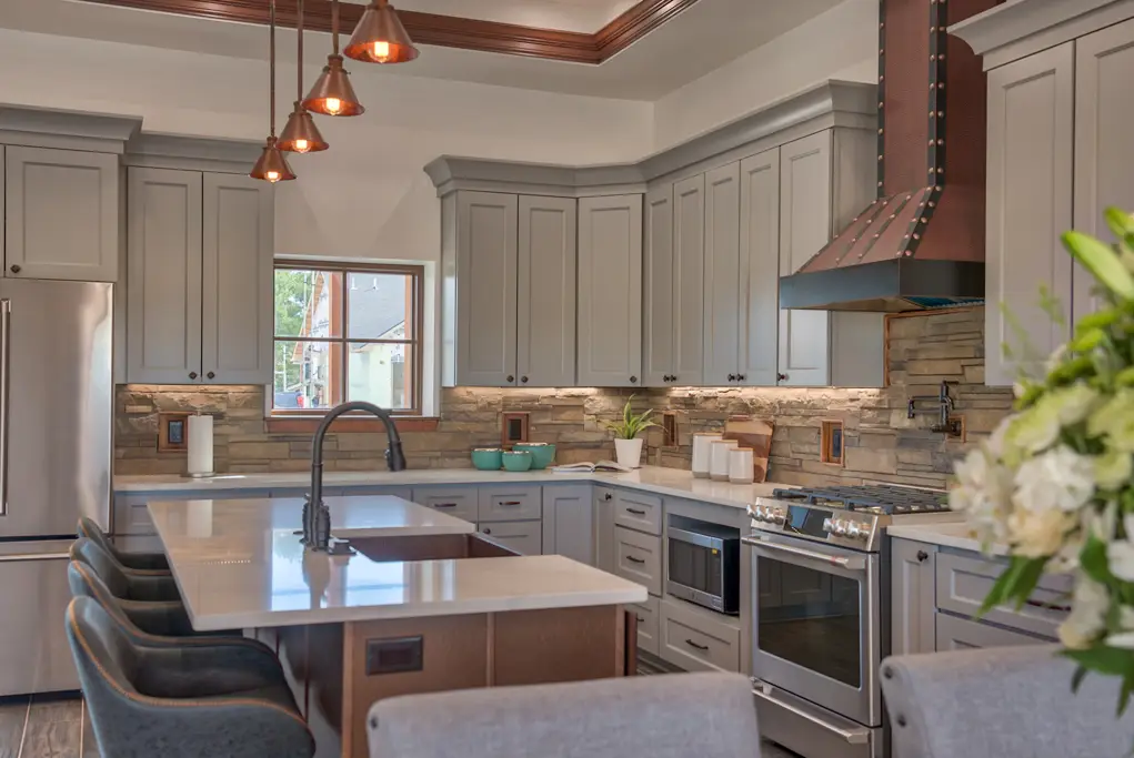 Interior photo of a furnished home built by Harvell Homes in Powell, Wyoming featuring a luxurious custom kitchen with tray ceiling, premium appliances, wood island with premium countertop, wall-mount pot filler faucet, farmhouse kitchen sink, elevated stone backsplash, and kitchen hood