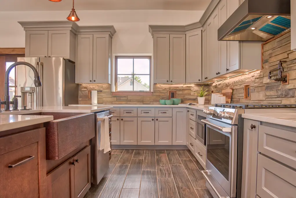Interior photo of a home built by Harvell Homes in Powell, Wyoming featuring a luxurious custom kitchen with tray ceiling, premium appliances, wood island with premium countertop, wall-mount pot filler faucet, farmhouse kitchen sink, elevated stone backsplash, kitchen hood and wood-look plank tile flooring