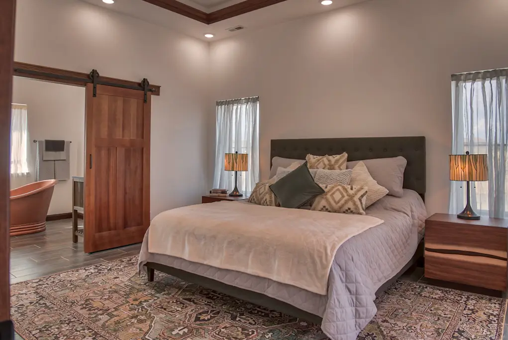 Interior photo of a home built by Harvell Homes in Powell, Wyoming featuring a luxurious and furnished bedroom with tray ceiling, sliding bathroom barn door leading to a bathroom with freestanding tub and custom sinks