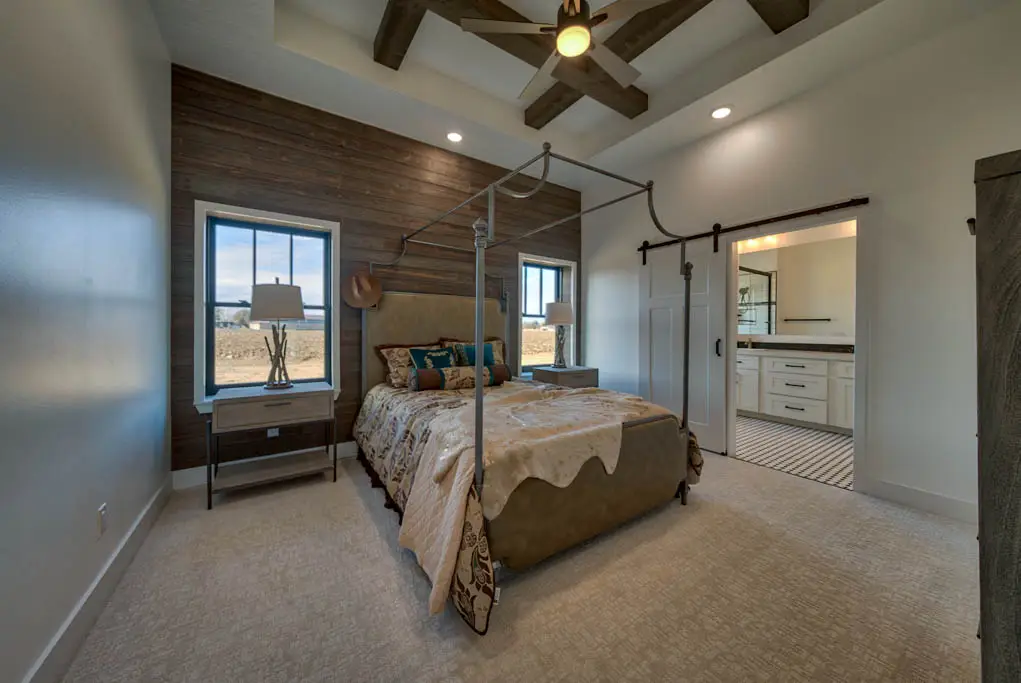 Interior photo of a home built by Harvell Homes in Powell, Wyoming featuring a luxurious and furnished master bedroom with tray and wood-beam ceiling, floor-to-ceiling shiplap statement wall, and sliding barn door to the bathroom