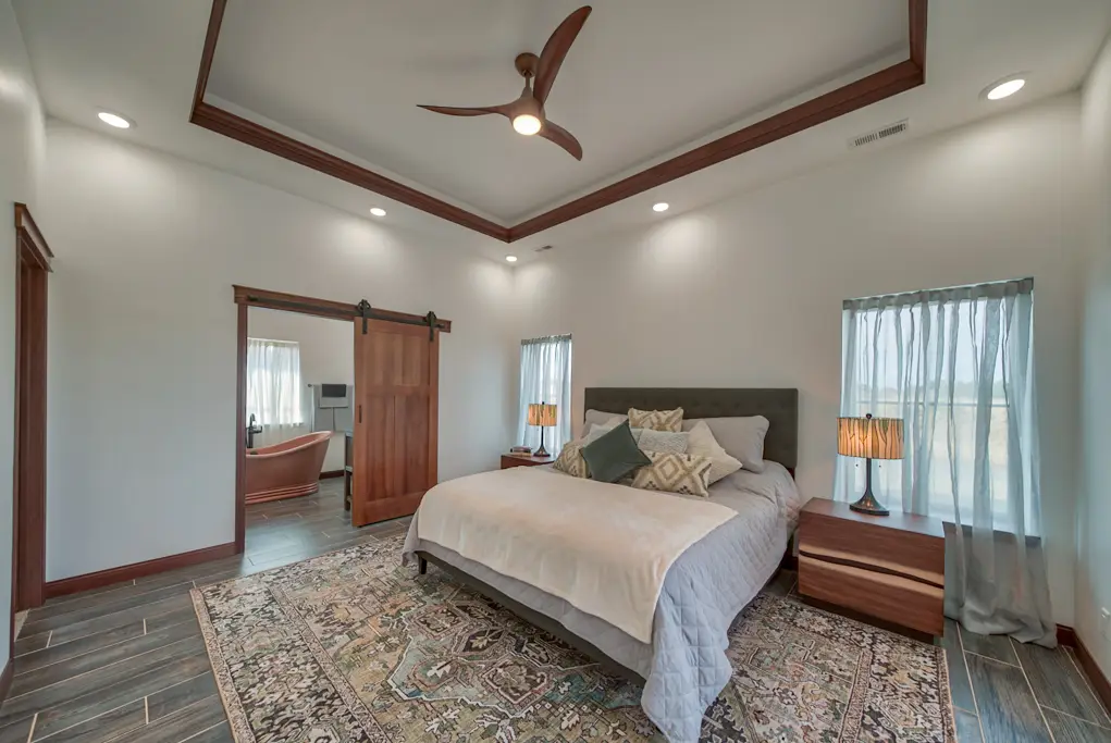 Interior photo of a home built by Harvell Homes in Powell, Wyoming featuring a luxurious and furnished bedroom with tray ceiling, sliding bathroom barn door leading to a bathroom with freestanding tub and custom sinks